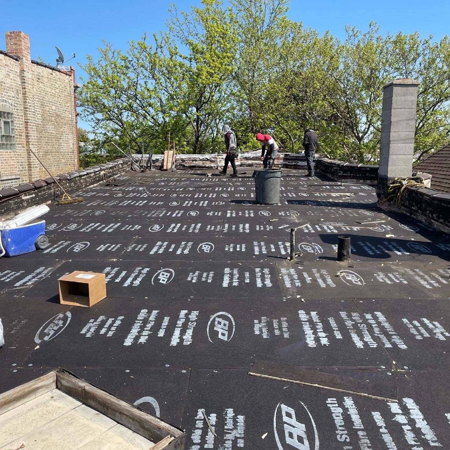Workers on a flat roof installing roofing material; trees and a brick building in background.