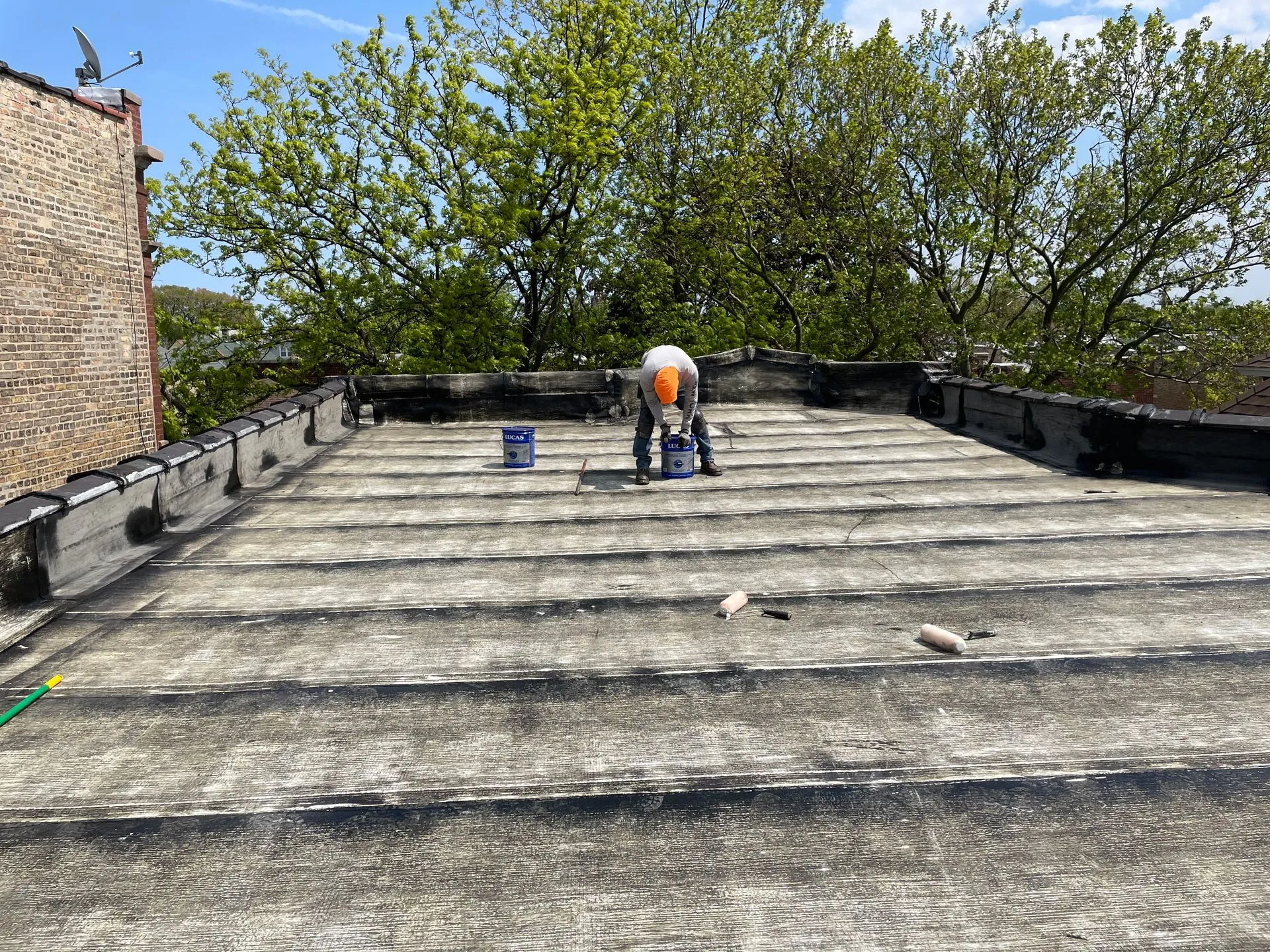 Person repairing a flat roof on a sunny day. Two paint cans nearby, green tree in background.
