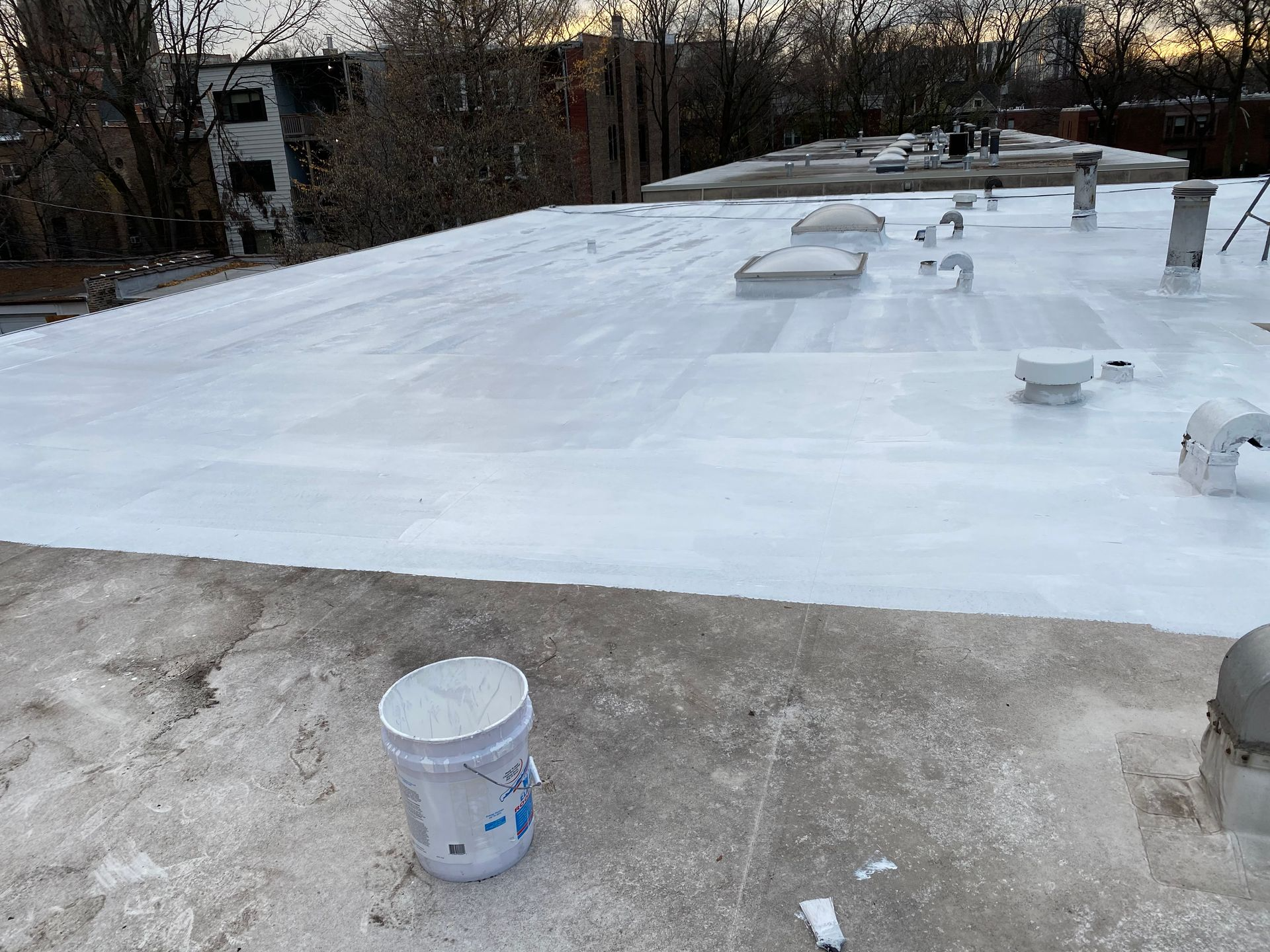 White-coated flat roof with vents and a bucket. Buildings and trees in the background. Overcast day.