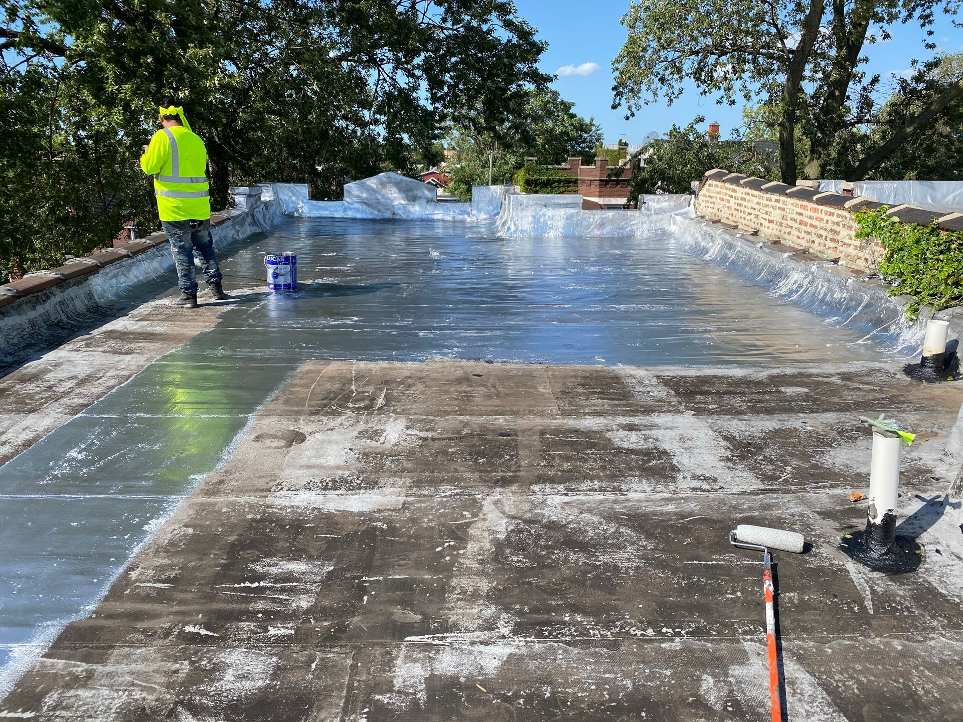 Person in safety vest on a flat roof, applying sealant. Roof partially coated, with blue bucket and roller.