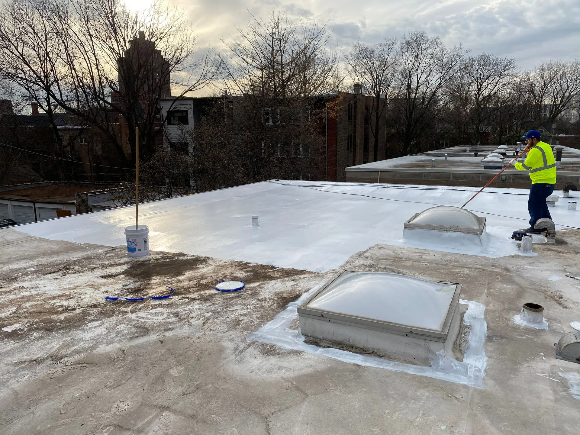 Person applying white sealant to a flat roof, near skylights, trees, and buildings.