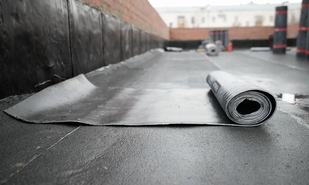 Roll of black roofing material being unrolled on a flat roof next to a brick wall.