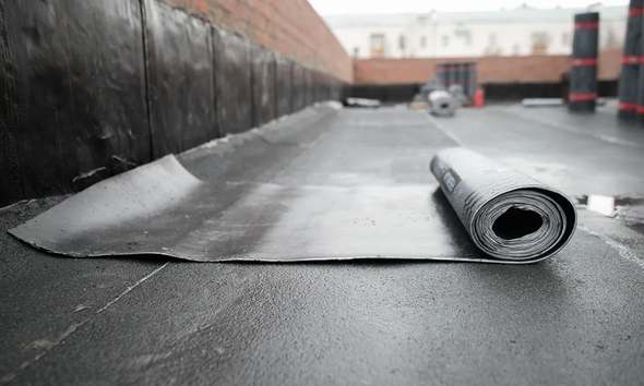 Roll of roofing material on a flat roof, near a brick wall and other rolls.