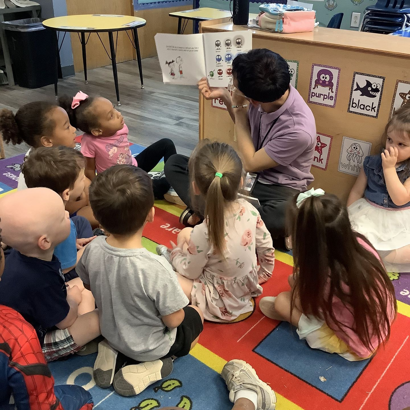 Children and an adult sit on a classroom carpet during story time near a bright window.
