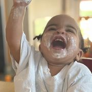 Laughing baby with messy white frosting, raising one arm indoors