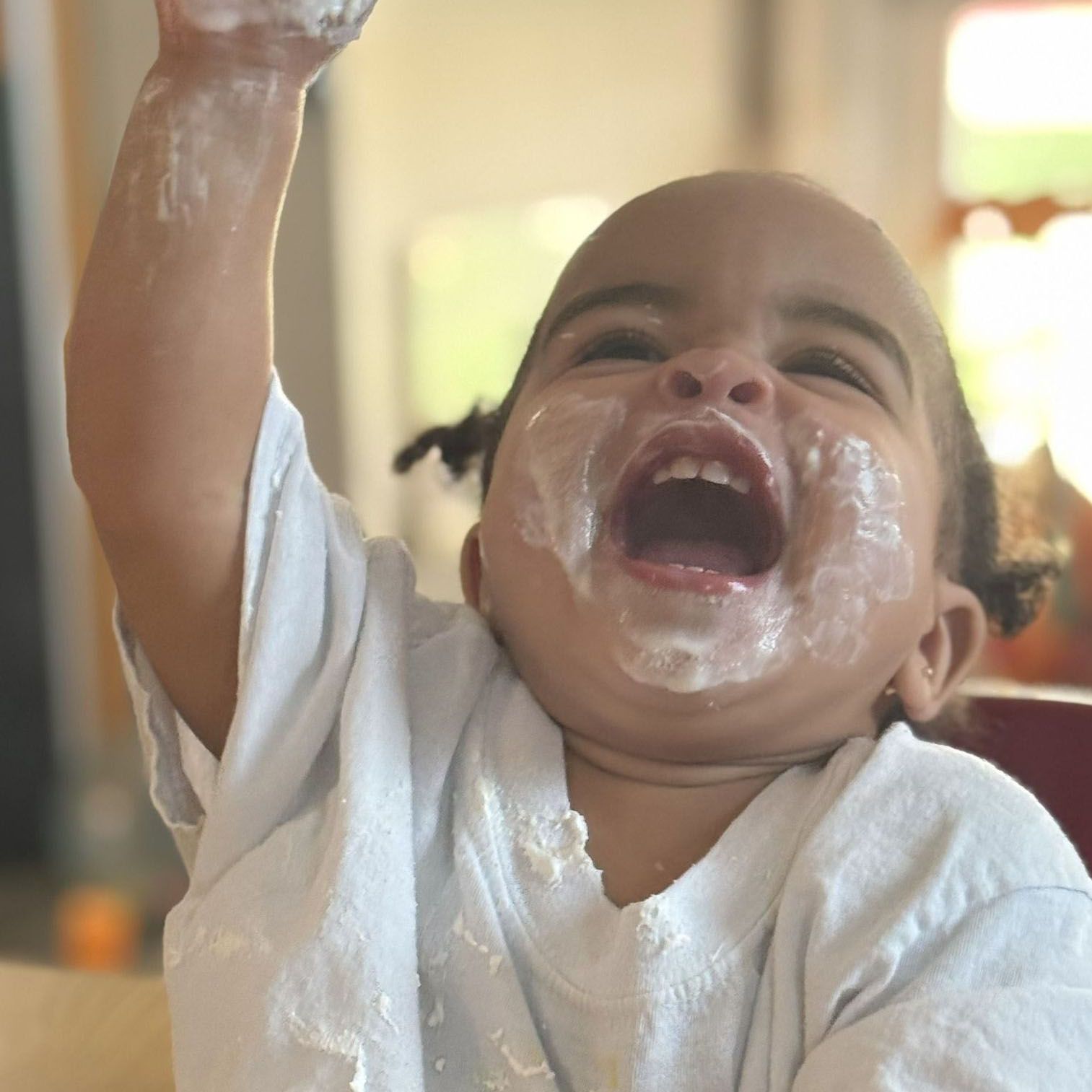 Laughing baby with messy white frosting, raising one arm indoors