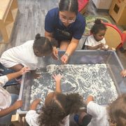 Children and a caregiver explore a sensory tray with white grains on a classroom floor