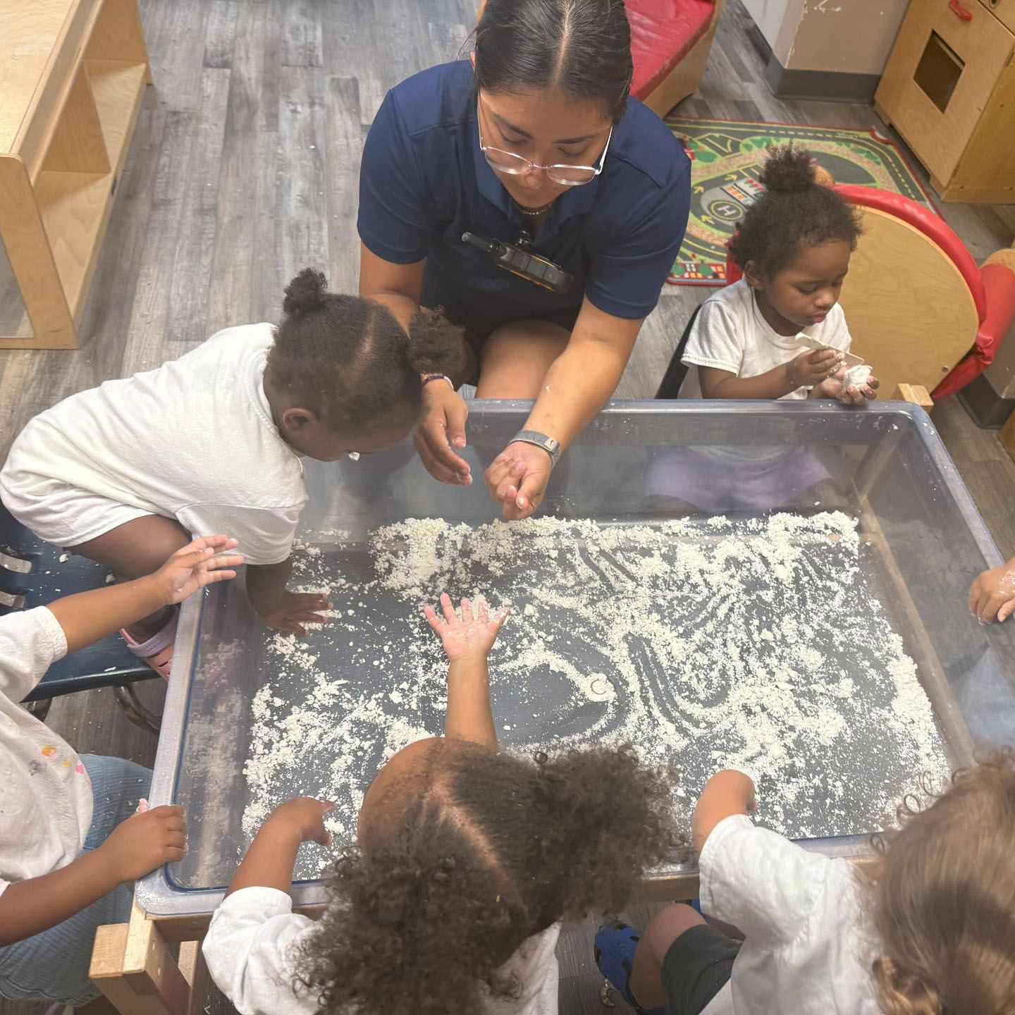 Children and a caregiver explore a sensory tray with white grains on a classroom floor