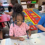 Child painting at a classroom table, focused on a white sheet with colorful art supplies nearby.
