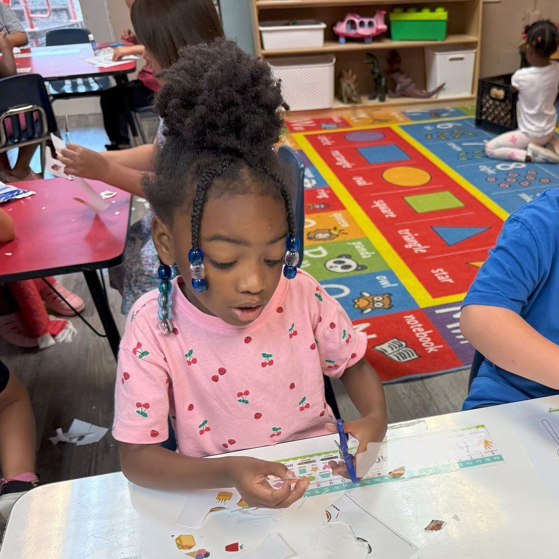 Child painting at a classroom table, focused on a white sheet with colorful art supplies nearby.