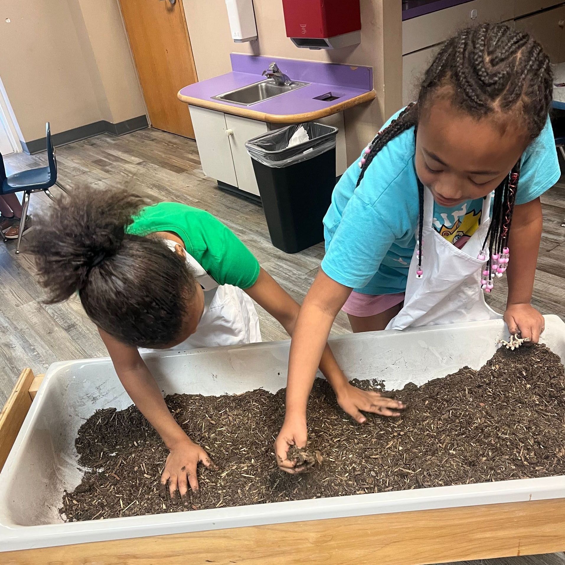 Two children in aprons digging in a sensory bin with soil in a classroom.