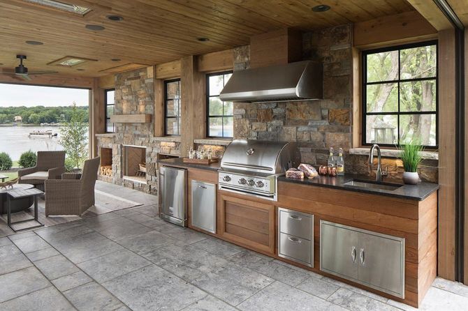 Outdoor kitchen with grill, stainless steel appliances, and stone wall, overlooking a lake.