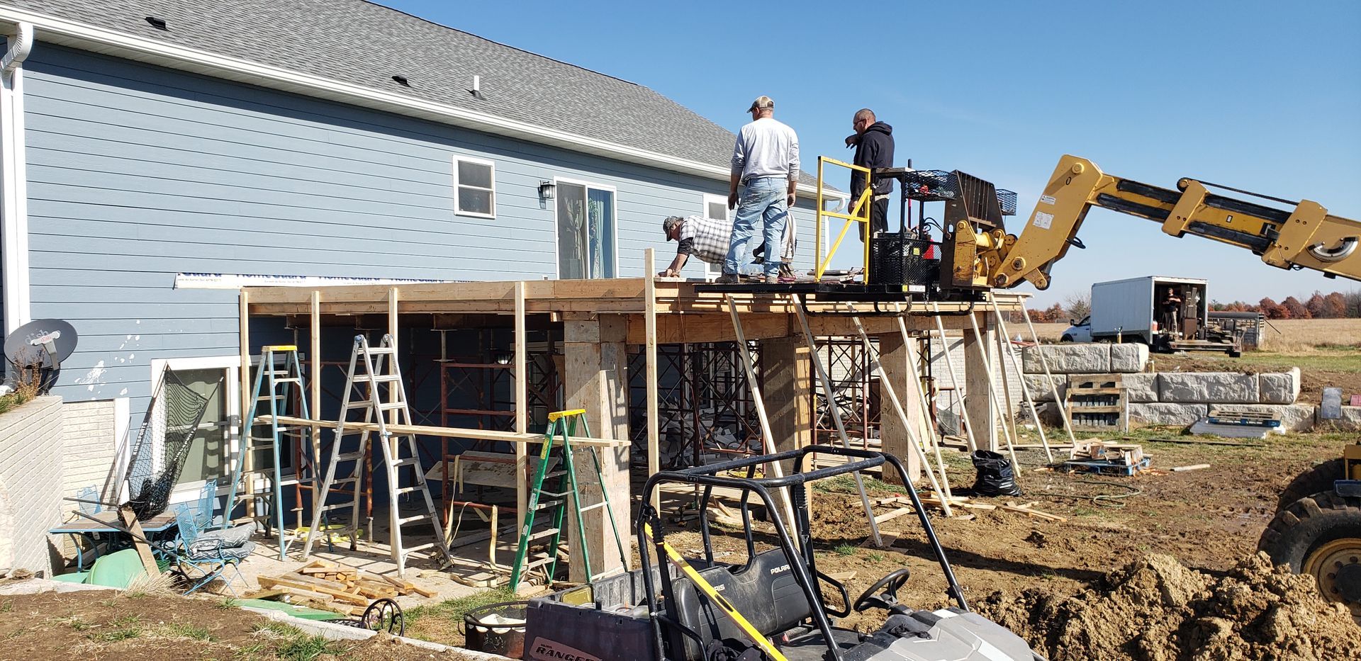 Construction workers build a porch extension on a house. An excavator is present.