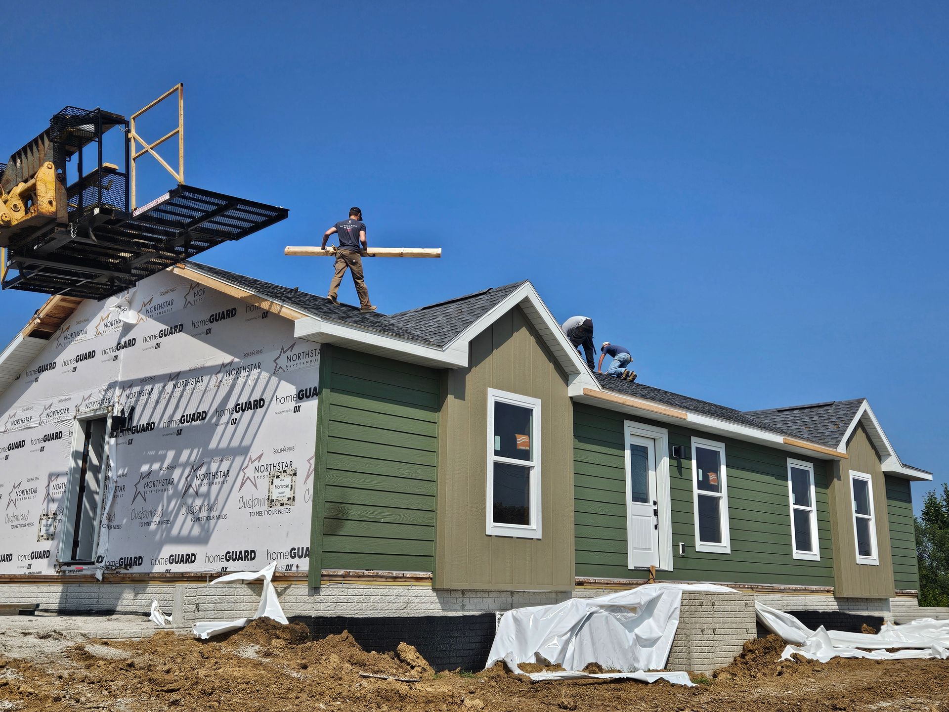 Workers building a house. Green siding, white wrap, blue sky. Crane with material.