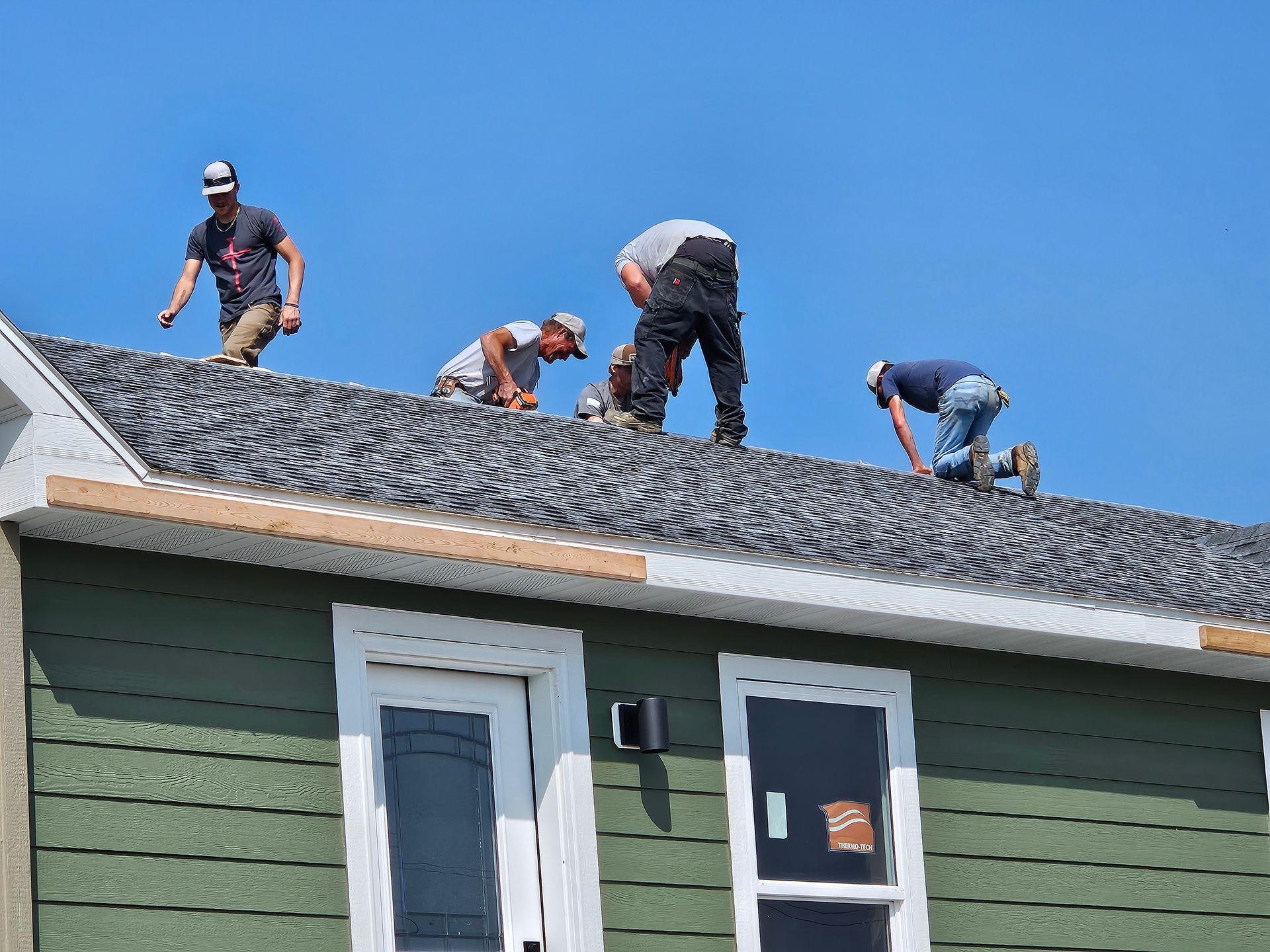 Construction workers on a roof, installing shingles, blue sky. Green siding below.