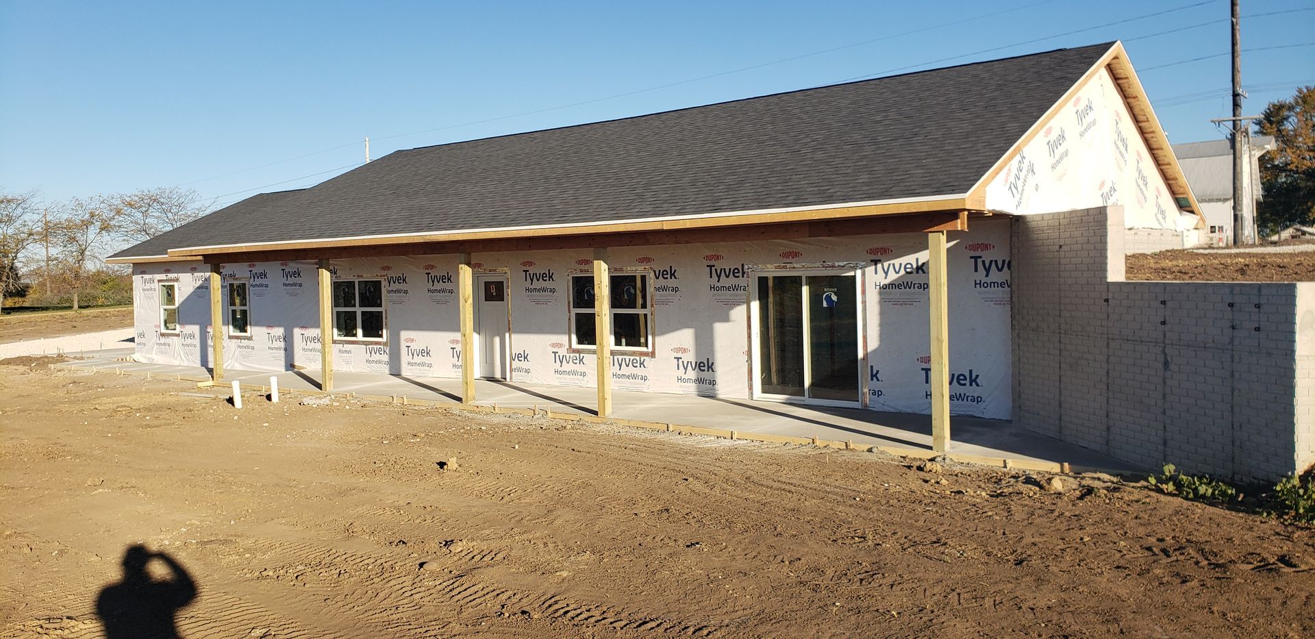 A building under construction with a black shingled roof, windows, and a porch on a sunny day.