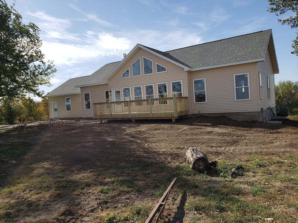 Beige house with gray roof, wooden deck, large windows, and unfinished landscaping on a sunny day.