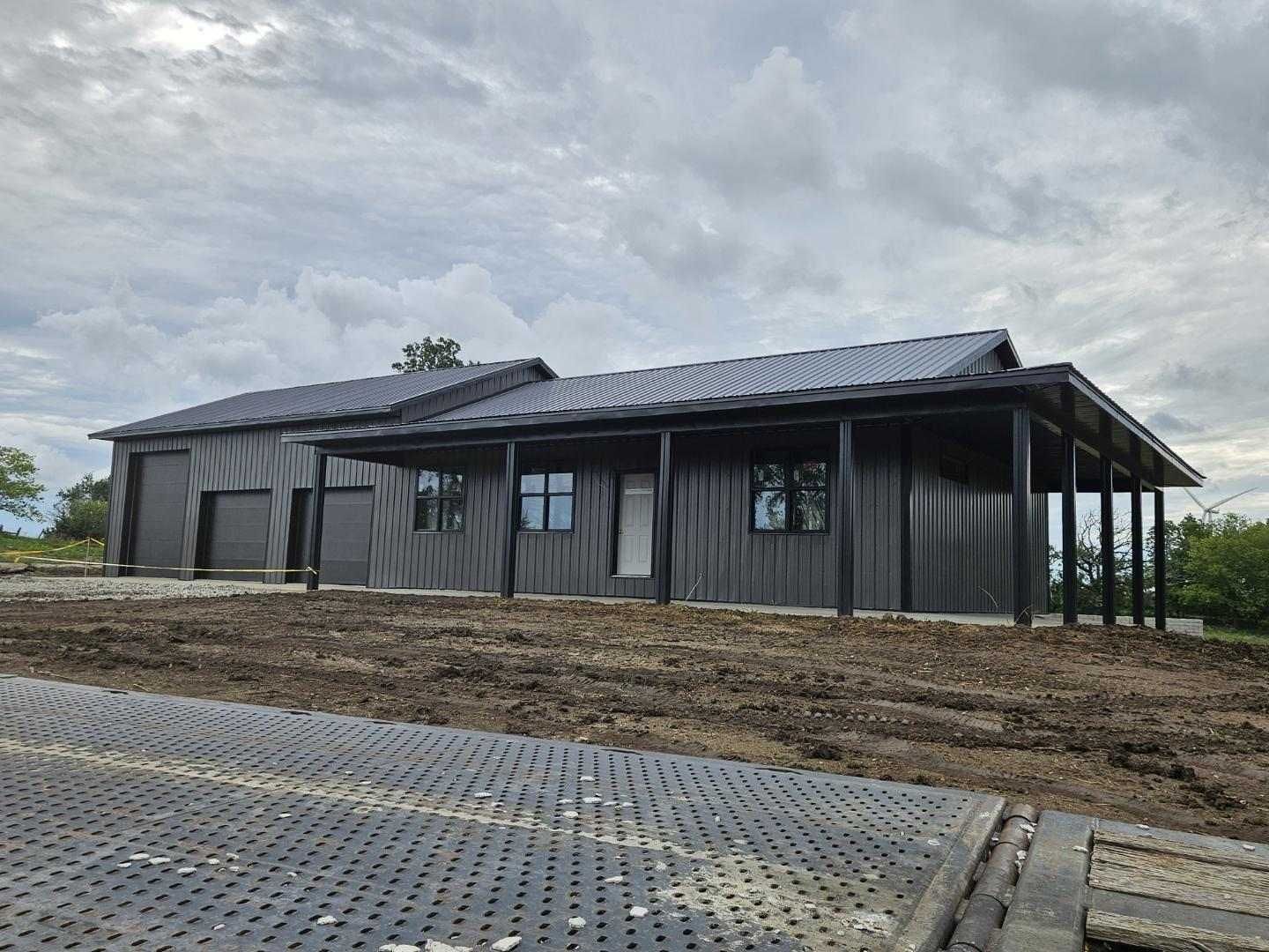 Black-sided house with covered porch and matching roof, under cloudy sky. Located on a dirt lot.