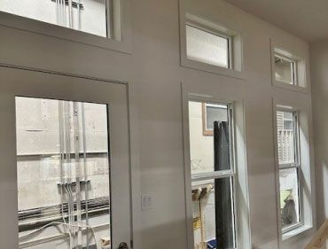 Interior wall with white-framed windows and a door. Natural light streams in; a partial view of the exterior is visible.