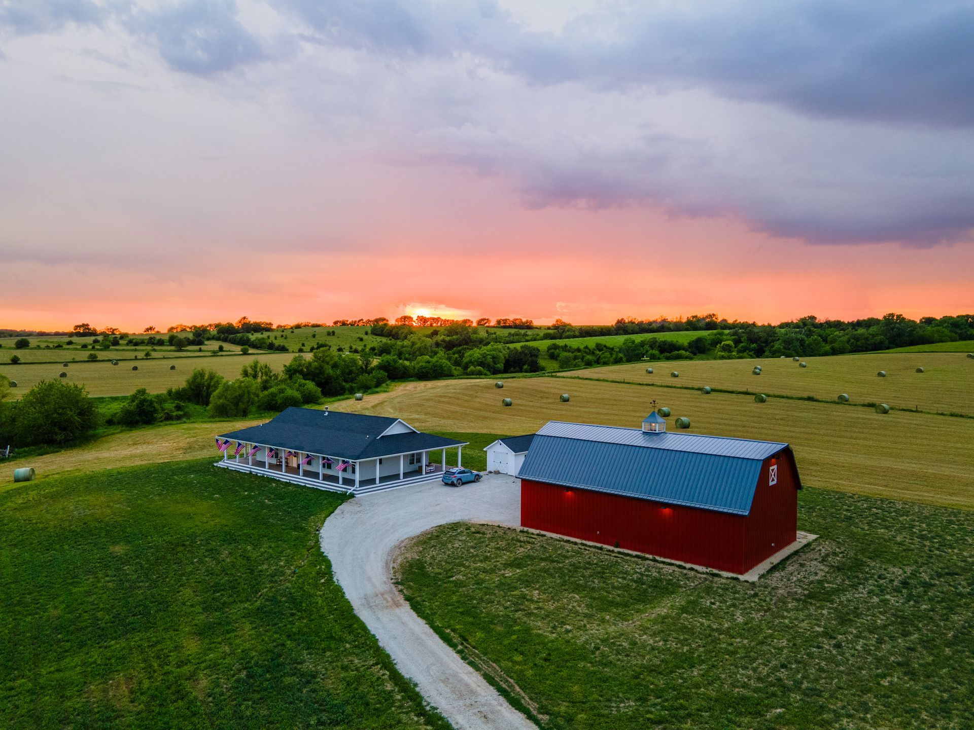 Red barn and house with a long driveway in a green field at sunset.