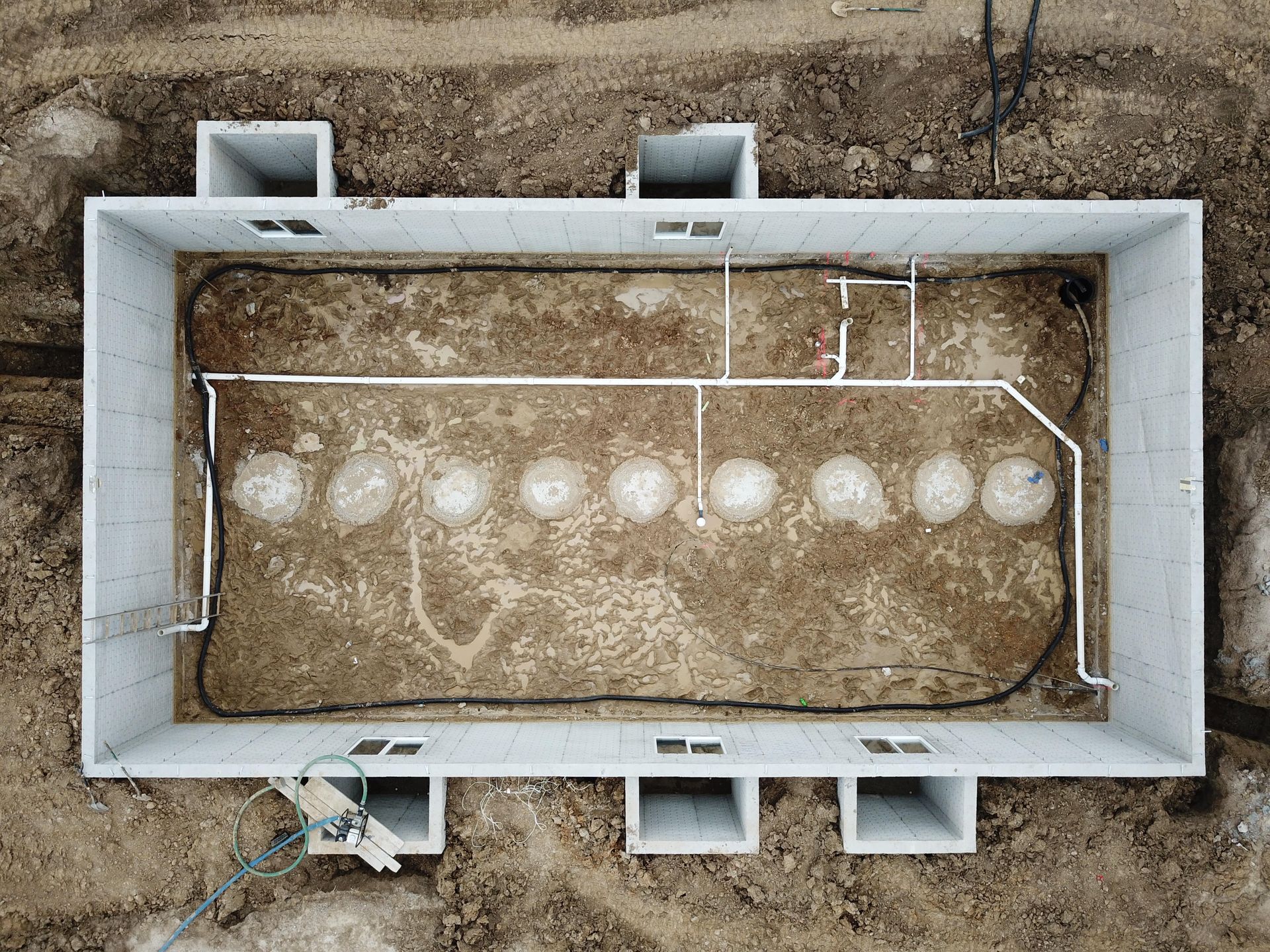 Aerial view of a rectangular concrete foundation with exposed soil and white plumbing, and several circular mounds.