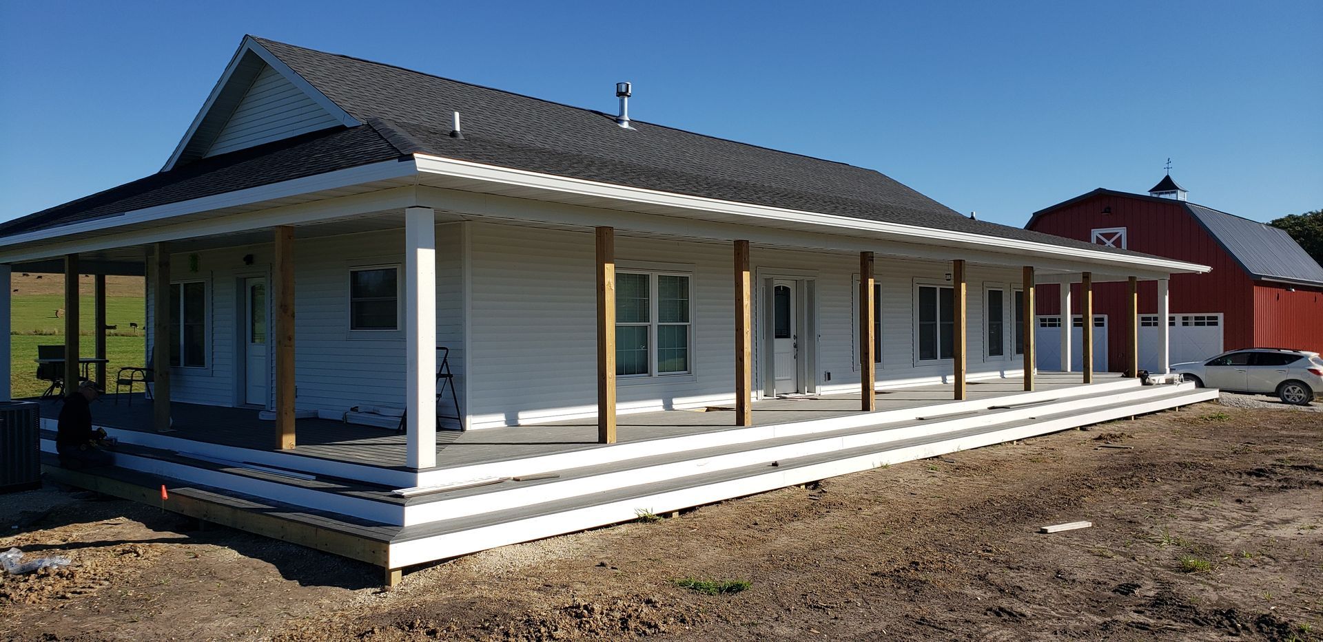 White house with wrap-around porch and a red barn on a sunny day.