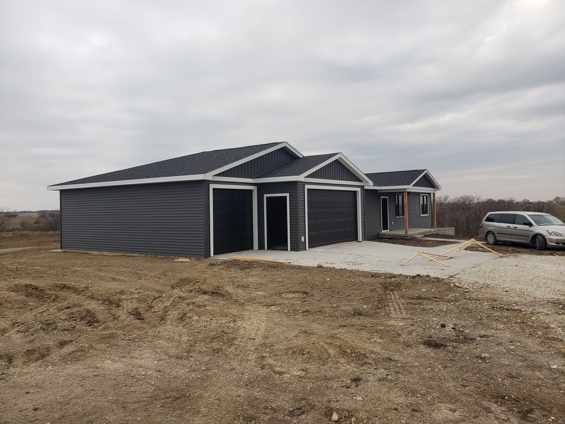 New house with dark siding and a two-car garage on a gravel lot under a cloudy sky.