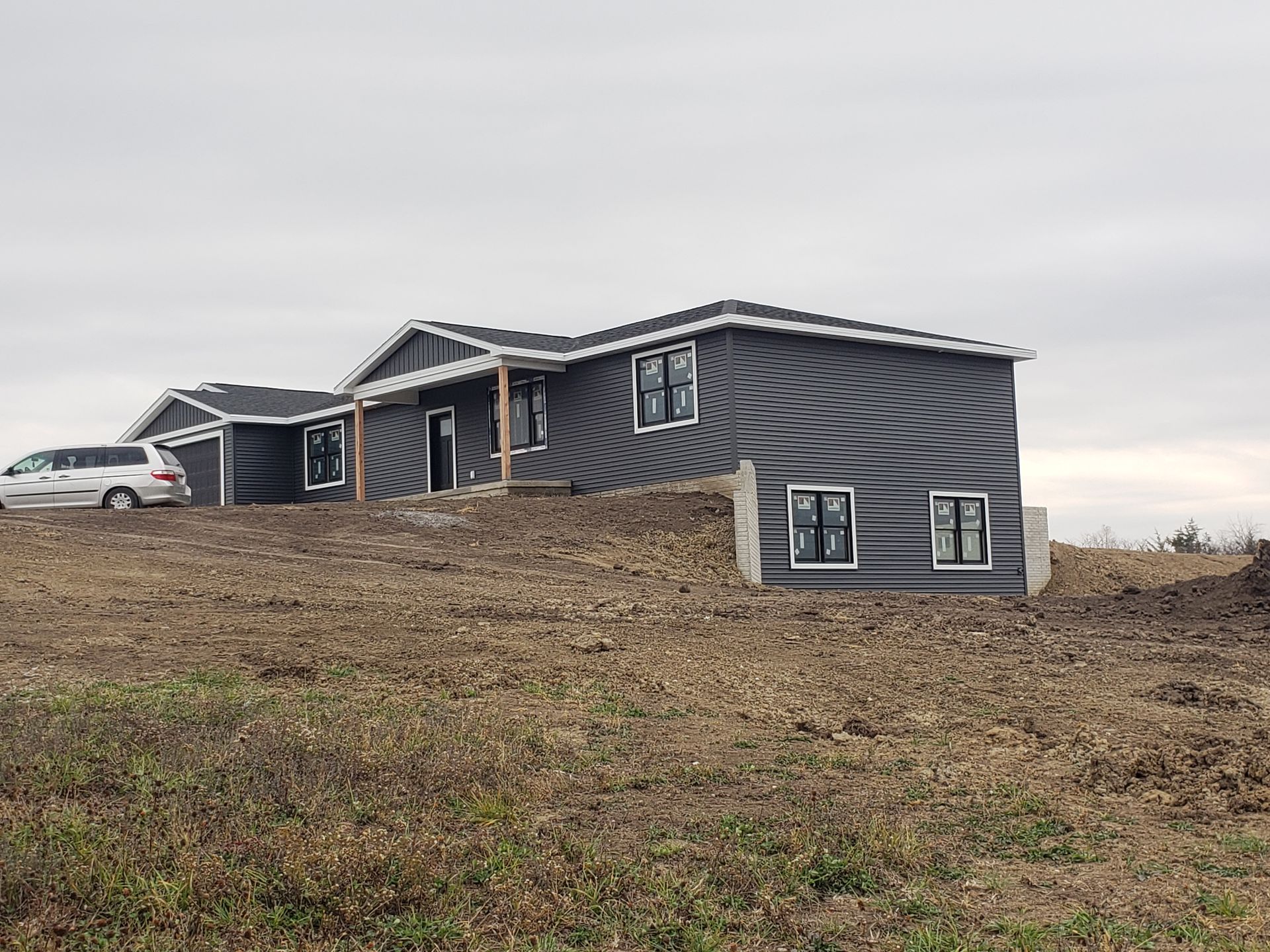 Newly constructed house with dark gray siding, black framed windows, on a mound of dirt, overcast sky.