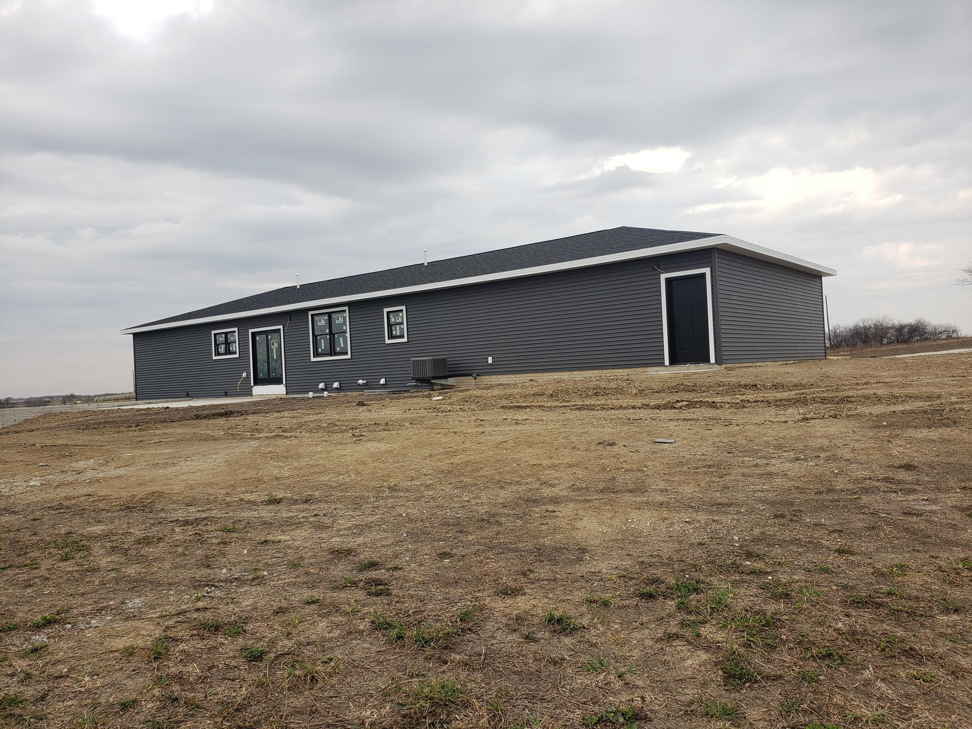 A gray house with a sloping roof stands on a brown field under a cloudy sky.