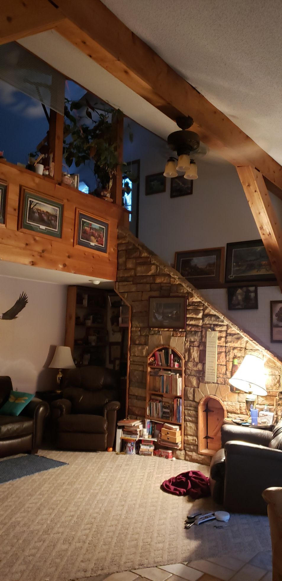 Cozy cabin interior with a loft, stone staircase, exposed beams, and multiple framed pictures on the walls.