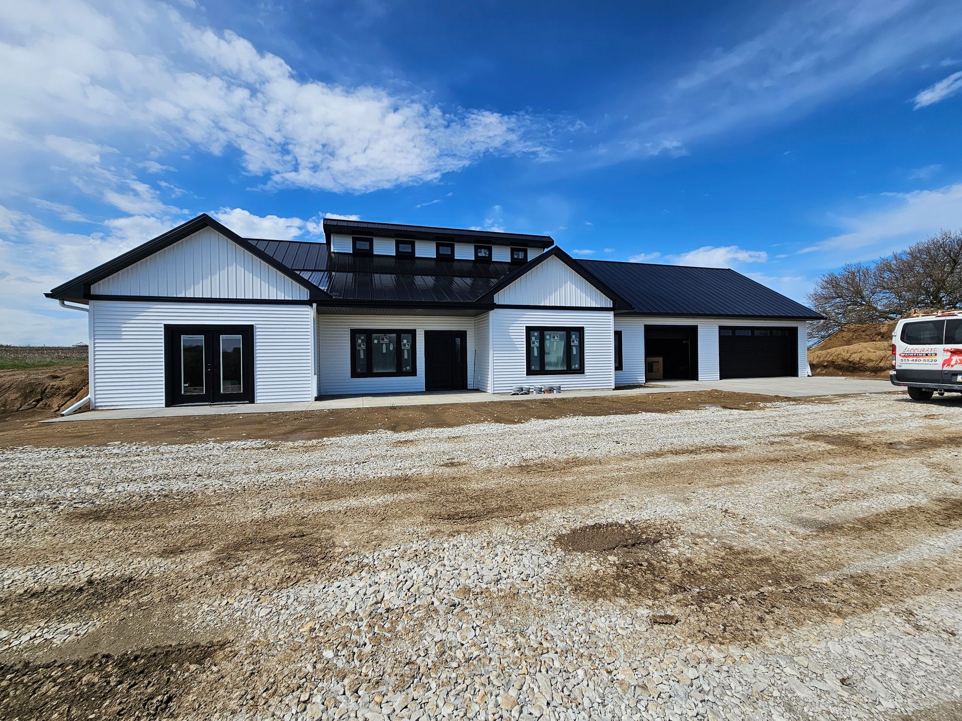 Modern white house with black trim, dark roof, and garage, on a gravel lot under a blue sky.