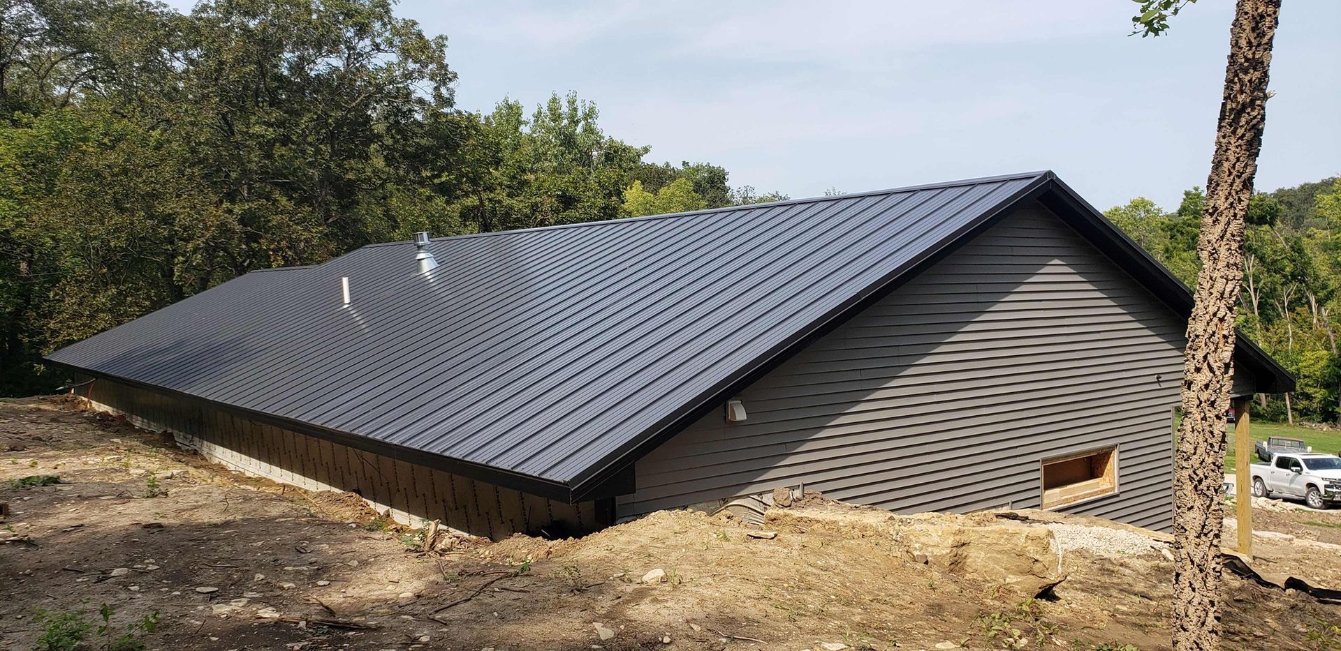 A modern, dark-roofed home under construction, surrounded by trees and a brown yard.