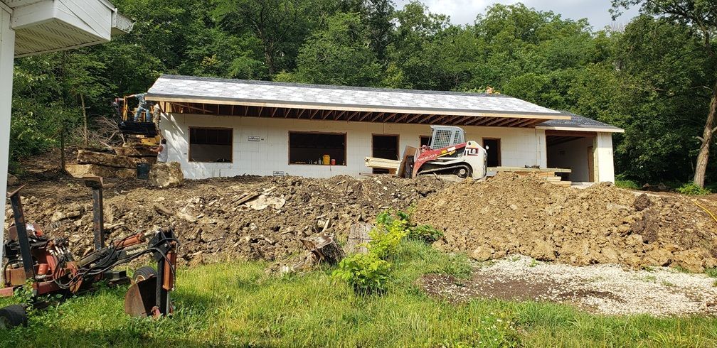 A house under construction with heavy machinery and piles of dirt. Green grass and trees in the background.