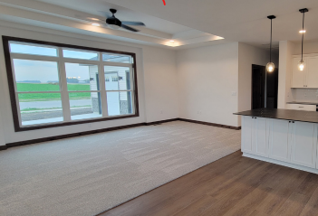 Empty, light-filled living room with large window, neutral carpet, and kitchen island.