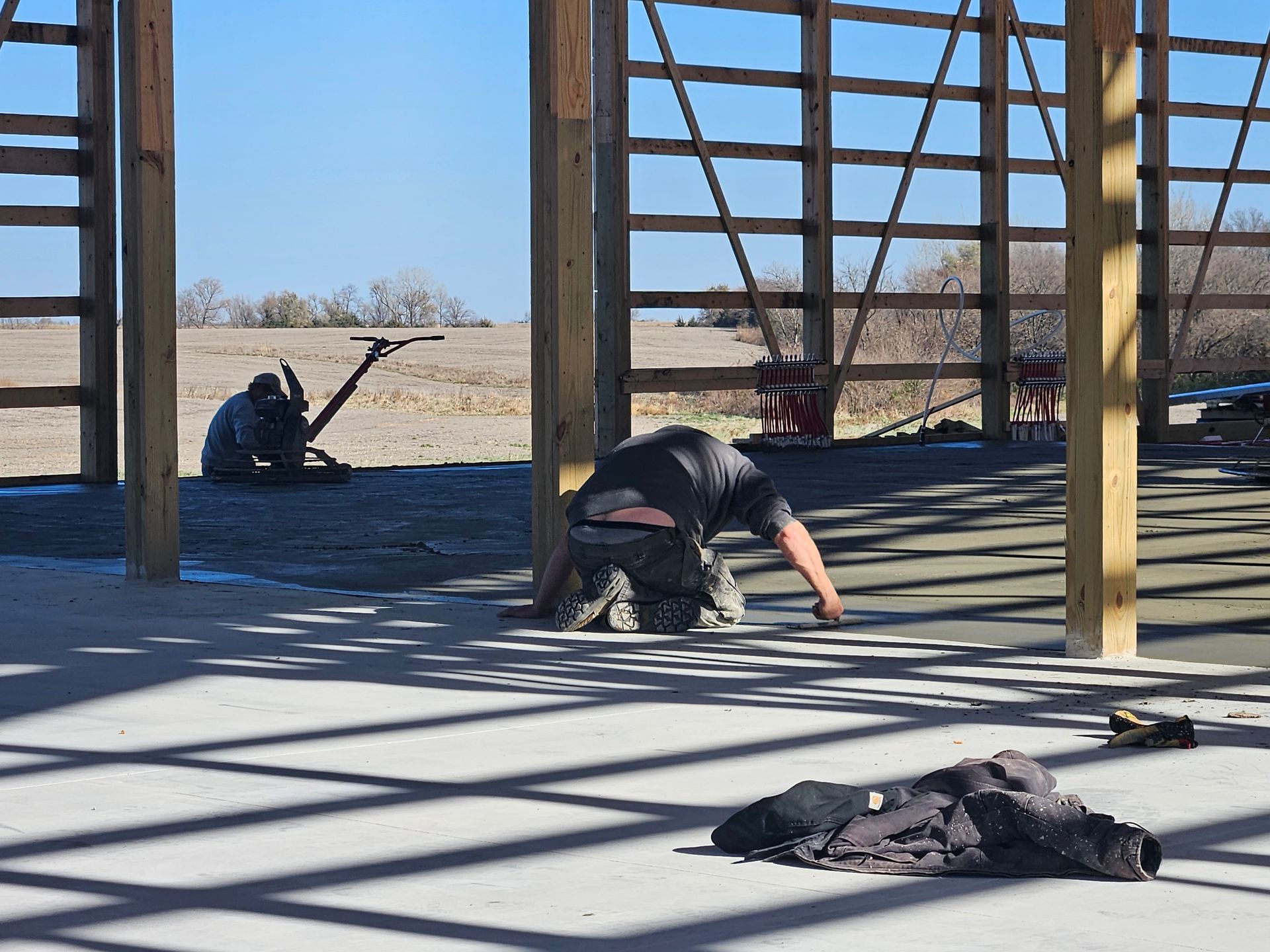 Two people working on a concrete floor inside a partially built wooden structure on a sunny day.