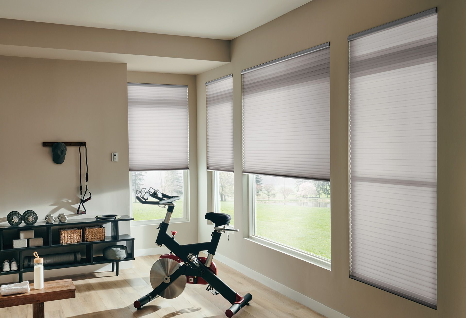 Home gym with spin bike, weights, and window shades. Beige walls, wood floor, natural light.