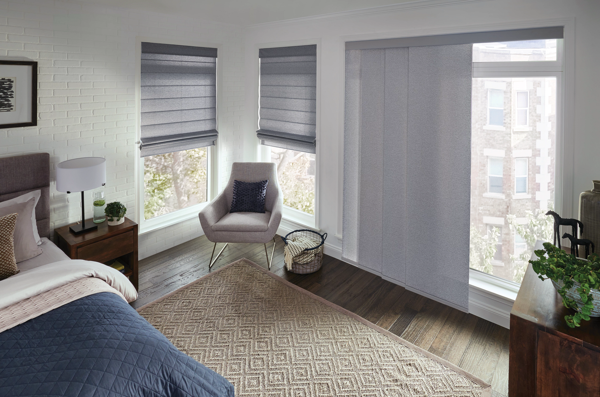Bedroom with gray window shades, rug, chair, and bed. Sunlight streams through the windows.