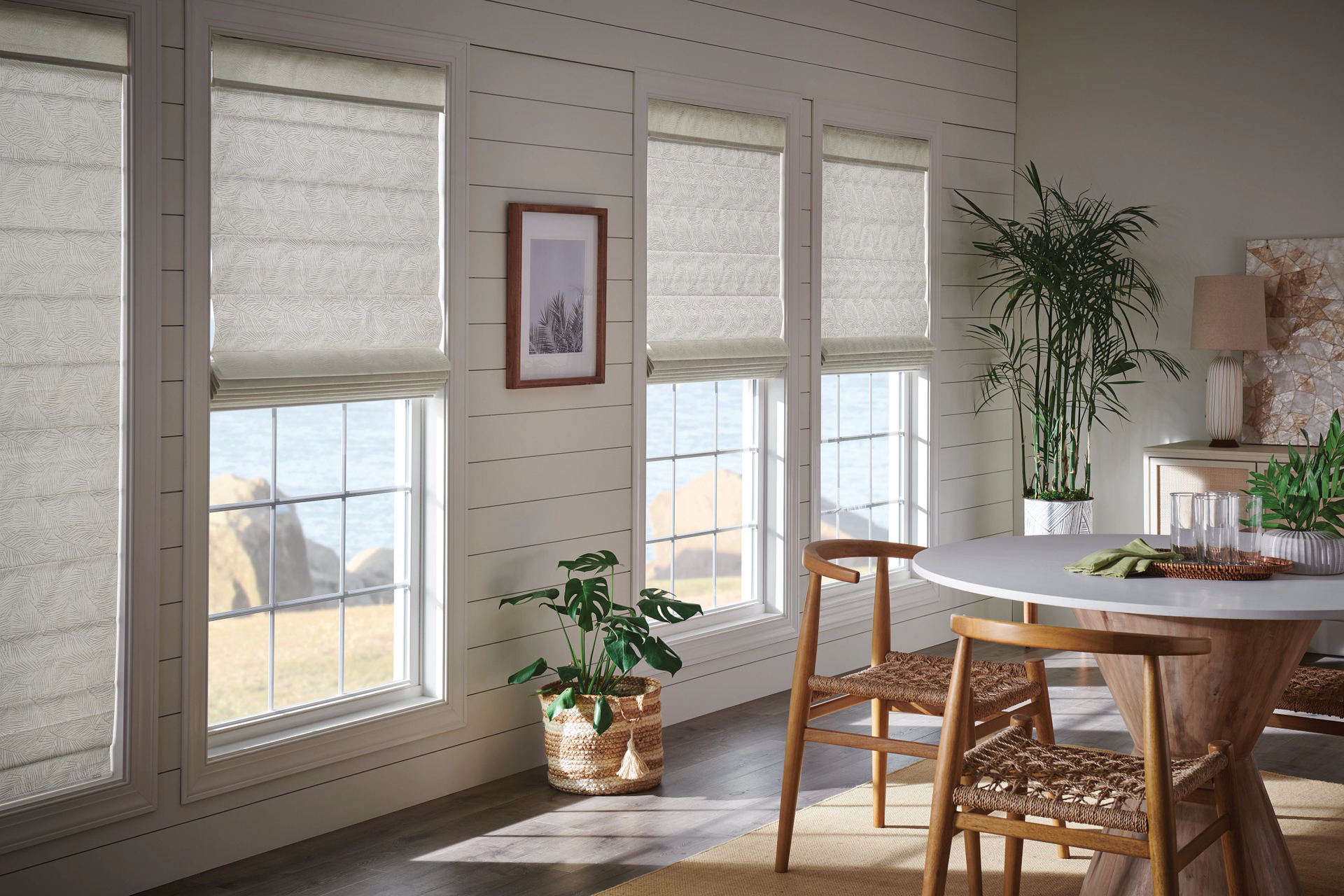 Bright room with windows, blinds, a table, chairs, plants, and a view of the water.