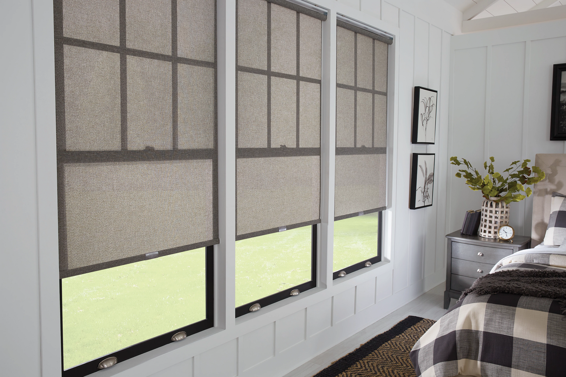 Three windows with beige roller shades in a white-walled room; a bed with a plaid blanket is visible.
