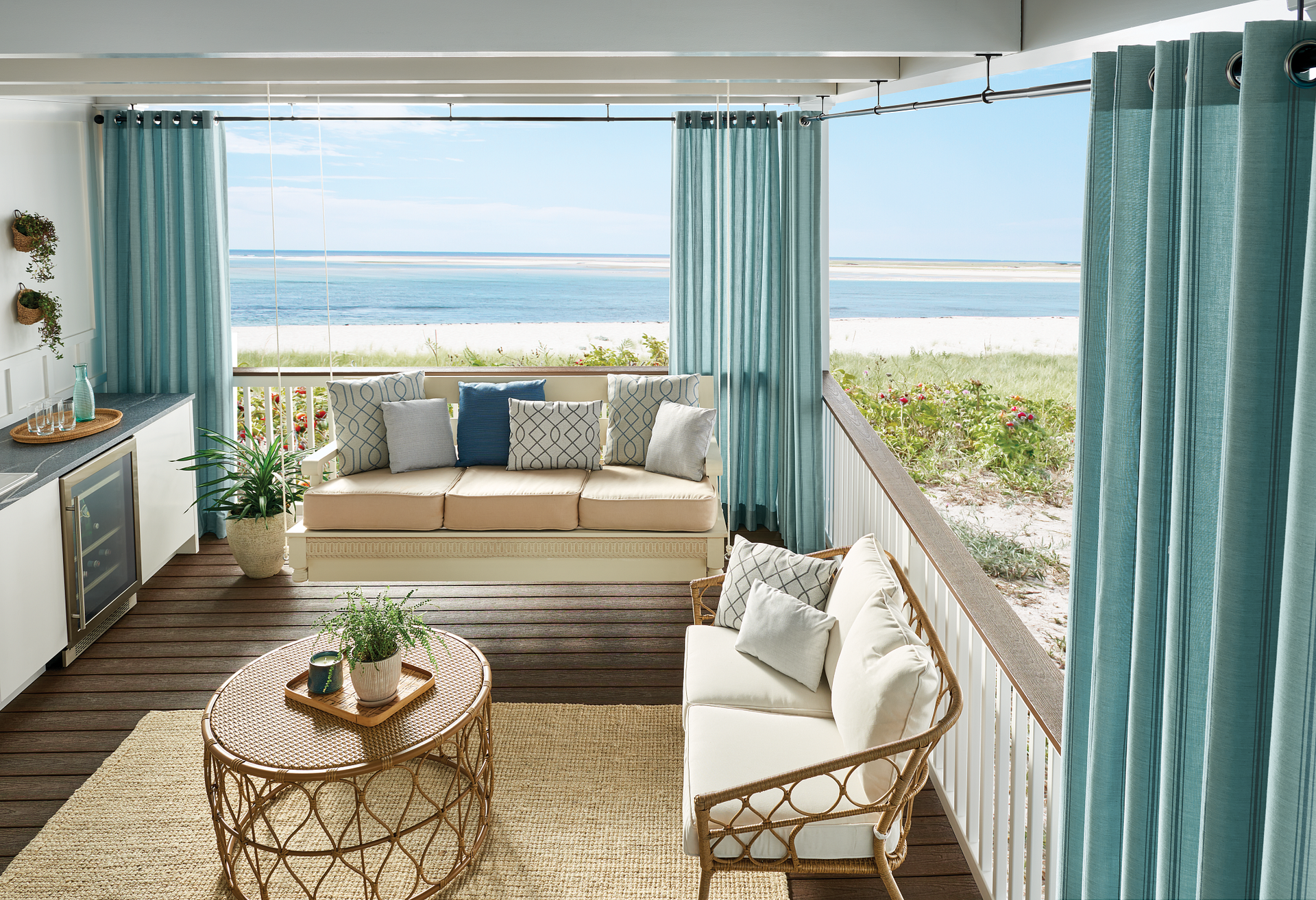 Beachside patio with aqua curtains, a tan couch, and wicker furniture, overlooking the ocean.