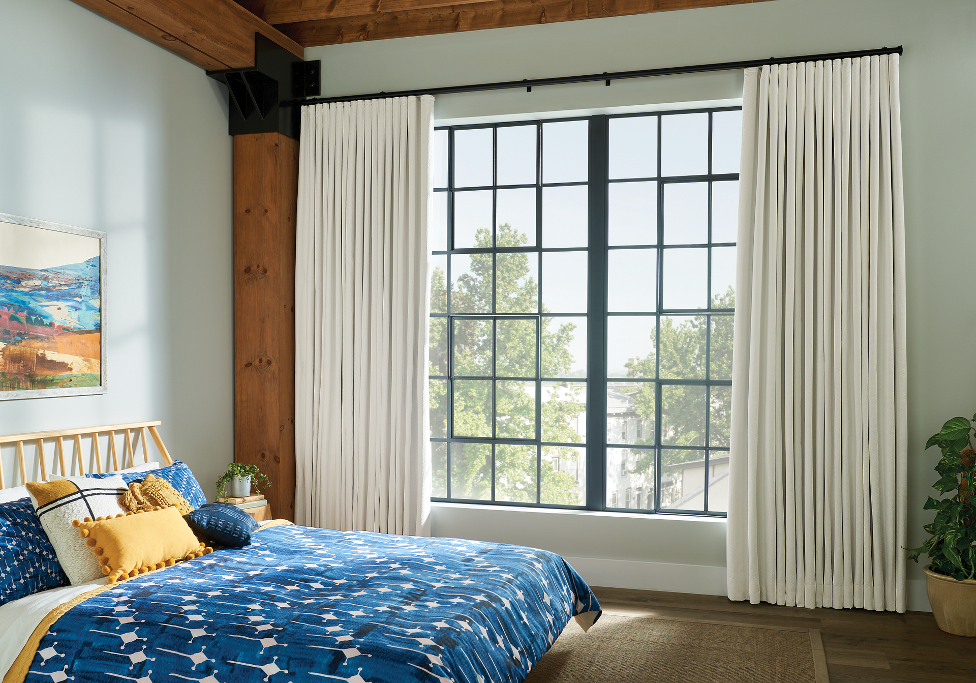 Bedroom with large window, cream curtains, blue bedding, wood bedframe, and a painting.