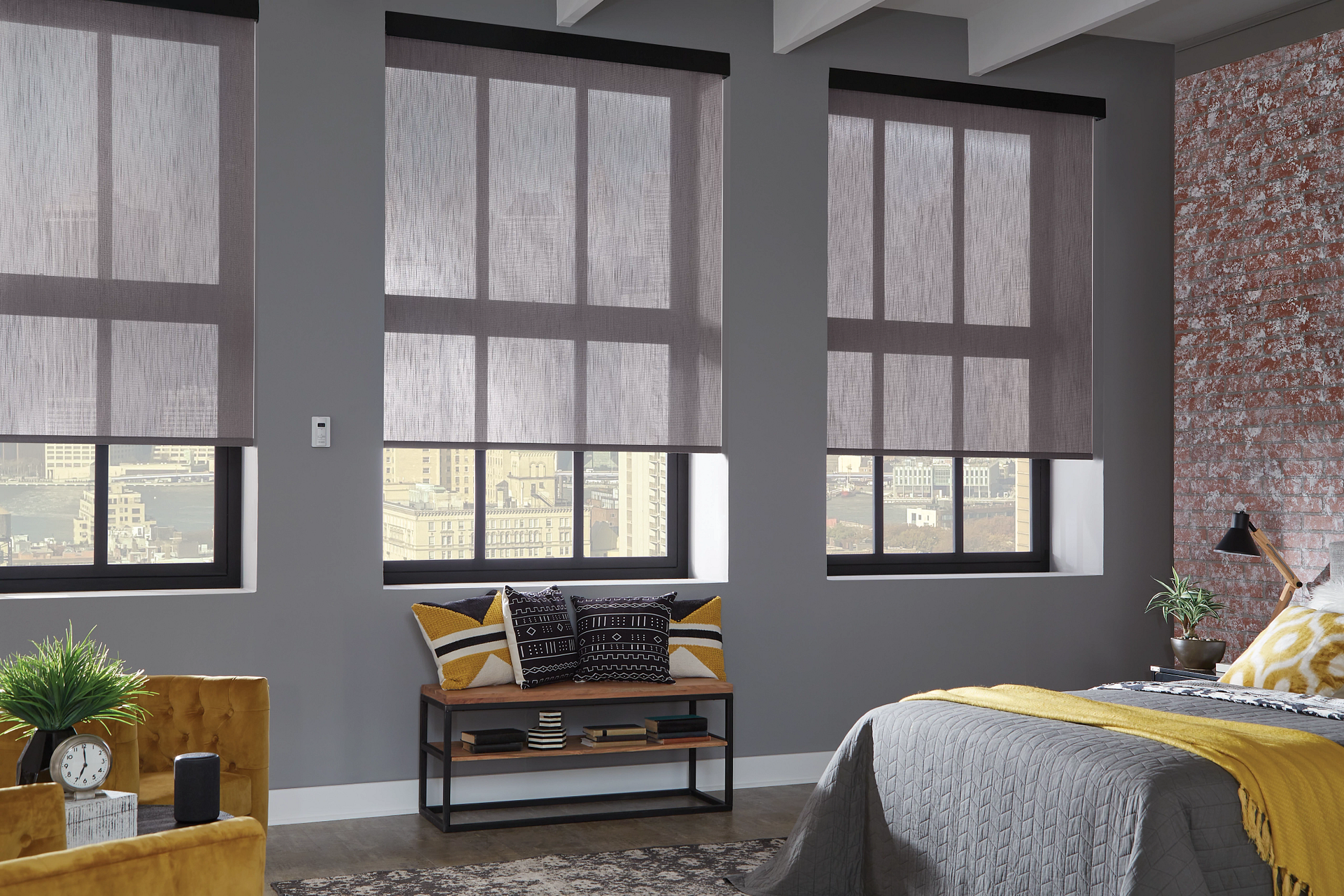 Bedroom with three windows, gray walls, and roll-up blinds. A bed with gray bedding and a yellow blanket is visible.