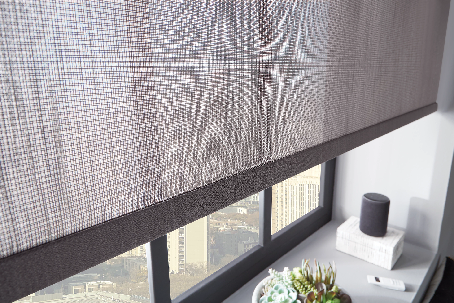 Close-up of a woven gray roller shade over a city view window, partially down. An Echo Dot sits on the windowsill.