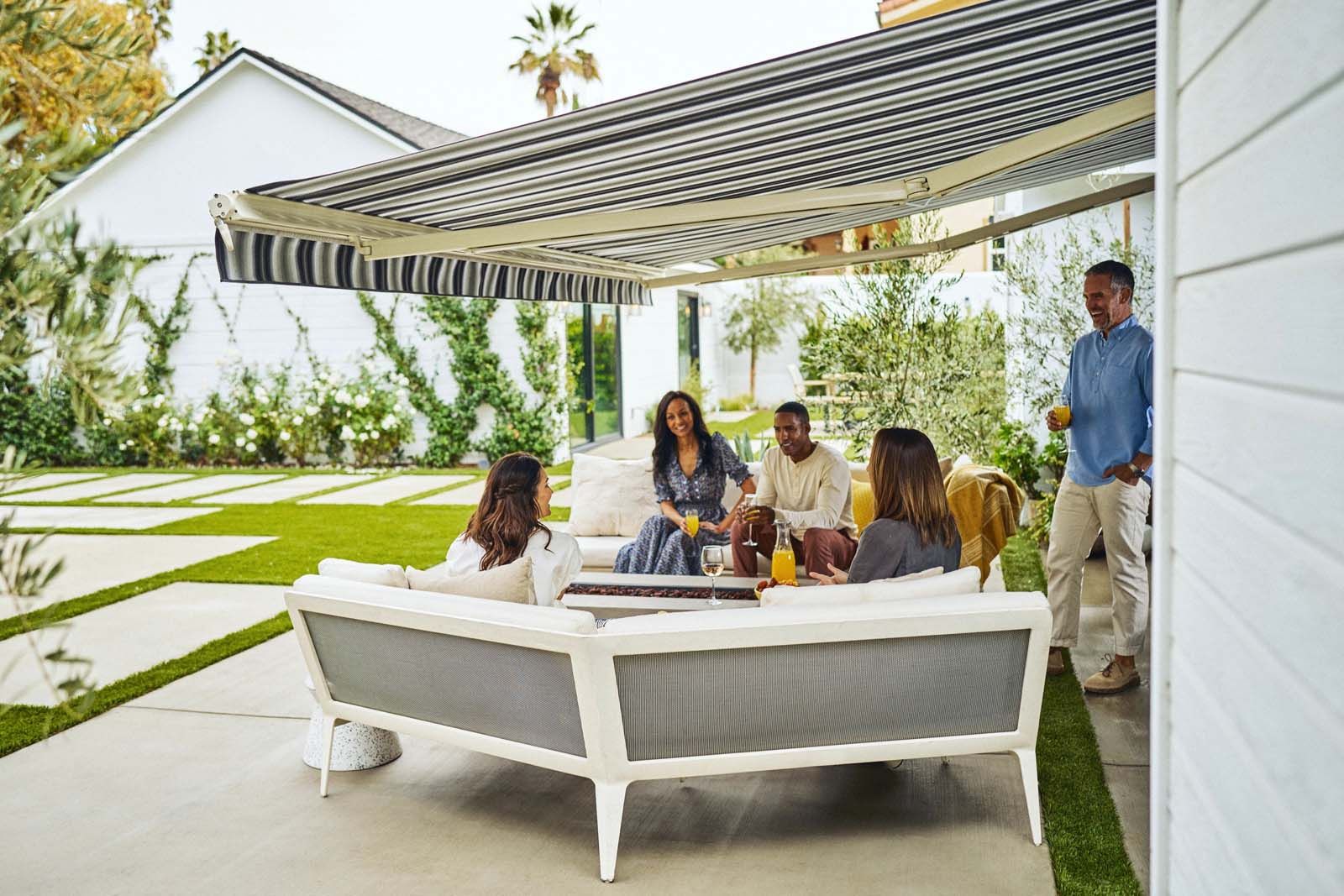 Five people relaxing on a white outdoor sofa under a striped awning in a yard.