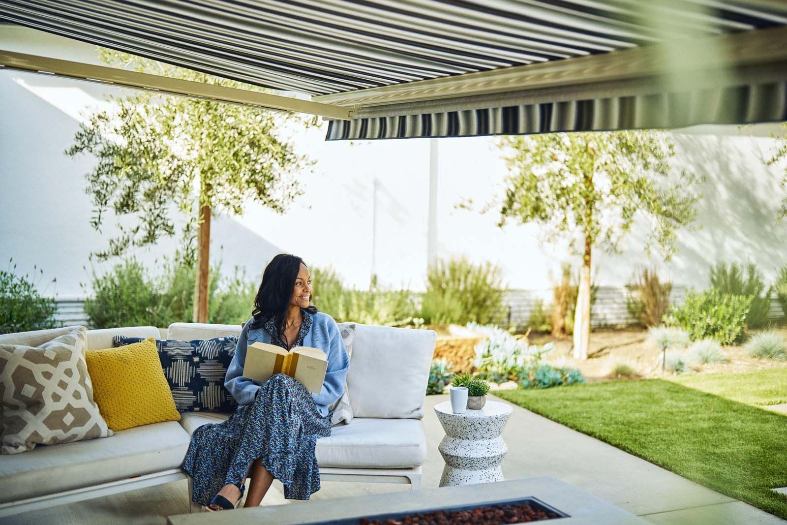 Woman in patterned dress on patio, holding gift, looking away, sunny outdoor setting.