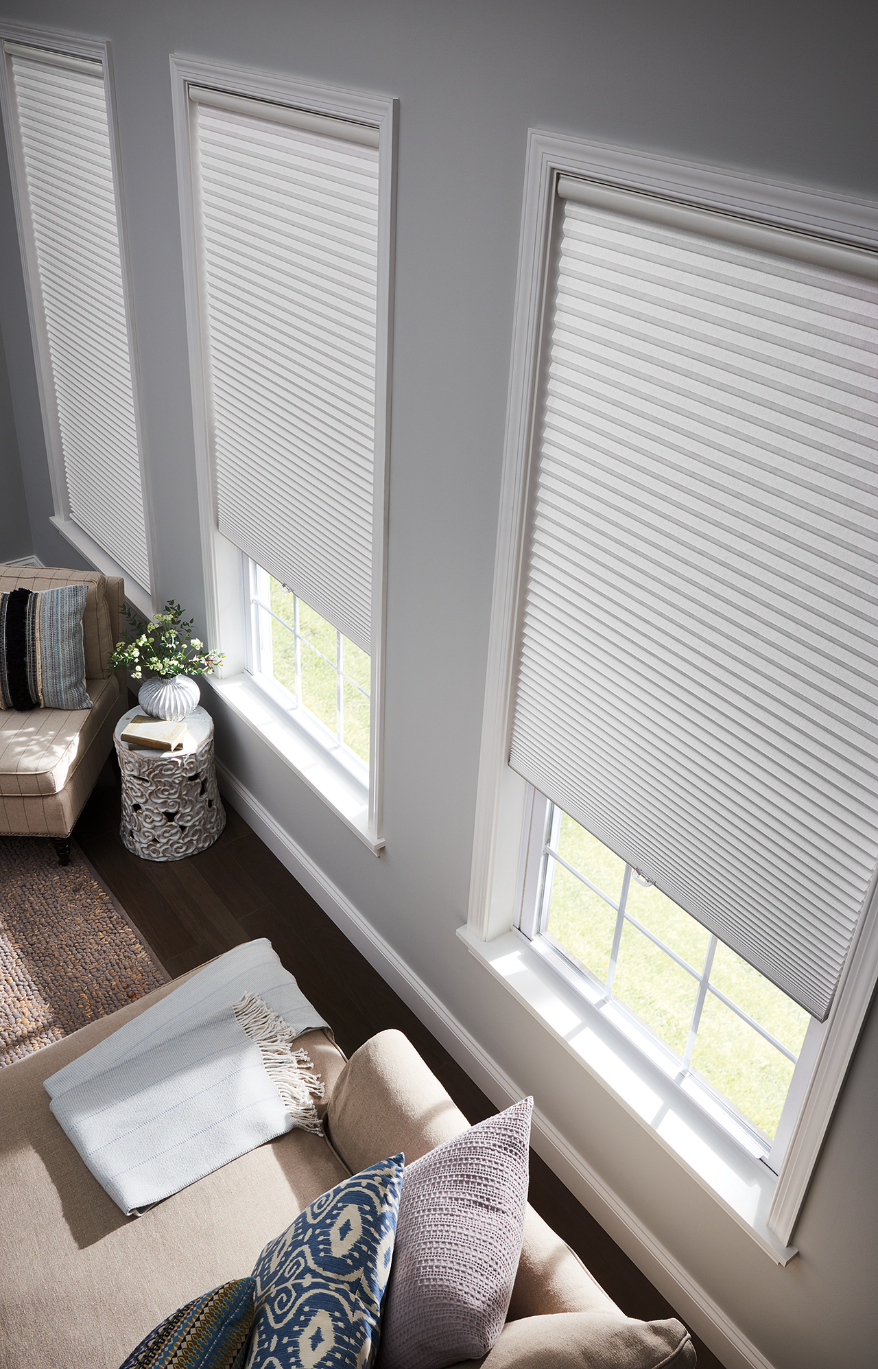 Three windows with white honeycomb blinds in a gray-walled room with furniture and area rug.