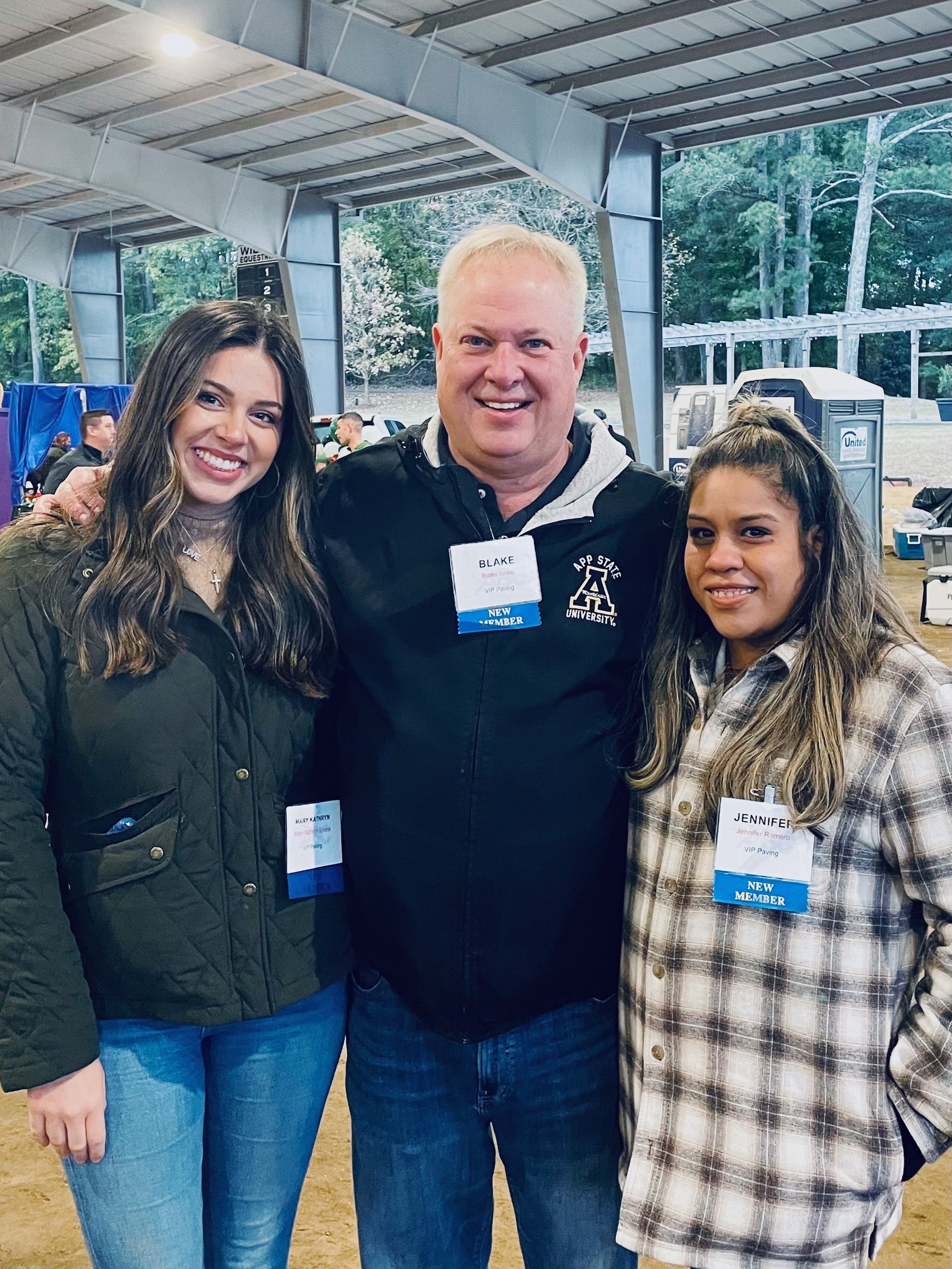Three people smiling, posing together outdoors under a shelter. Two women flank a man.