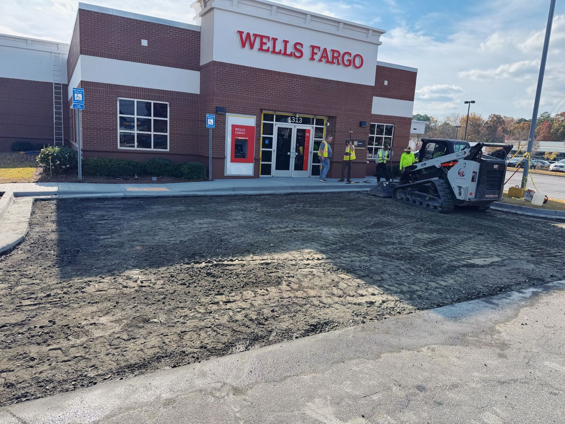 Construction in progress at a Wells Fargo bank. A skid steer is on freshly laid gravel. Workers are near the entrance.