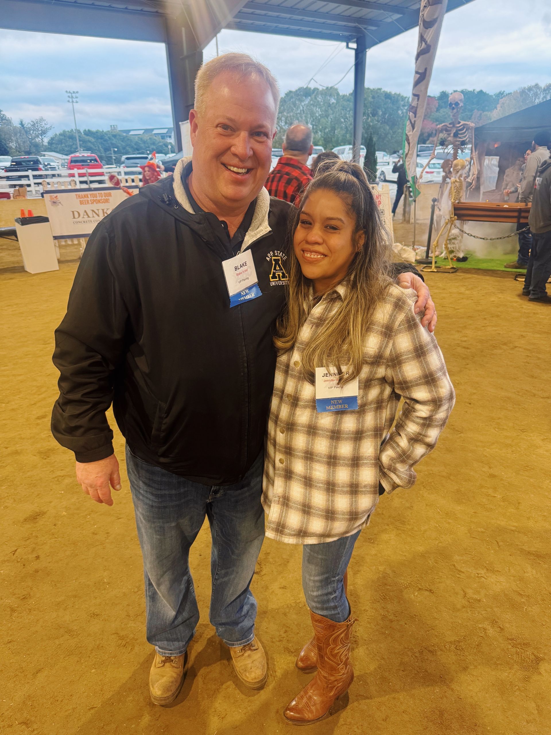 Two people smiling, posing together. Man in a black jacket and jeans. Woman in a plaid shirt and jeans. Outdoors.