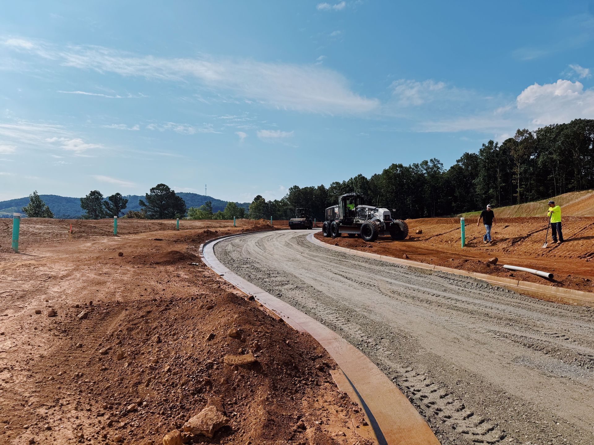 Construction site with graded road, heavy machinery, and workers. Clear sky and surrounding trees.
